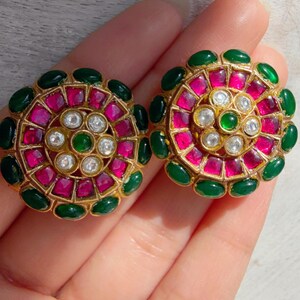 May include: Gold-toned stud earrings with a floral design. Each earring features a central cluster of clear stones surrounded by pink and green gemstones. The earrings are held in a hand.