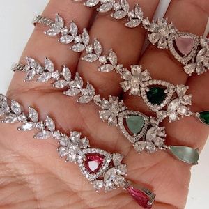 Może przedstawiać: Cztery srebrne kolczyki wiszące w kształcie liścia z kamieniami szlachetnymi w kształcie łzy. Kolczyki zdobią przezroczyste cyrkonie i różne kolorowe kamienie szlachetne, w tym różowy, zielony i jasnoniebieski.