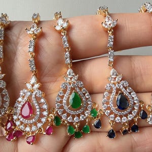 Może przedstawiać: Pięć złotych, ozdobnych kolczyków z wisiorkami w kształcie łezki. Każdy kolczyk ma centralny kamień szlachetny w kolorze różowym, czerwonym, zielonym, niebieskim i przezroczystym, otoczony mniejszymi, przezroczystymi kamieniami.