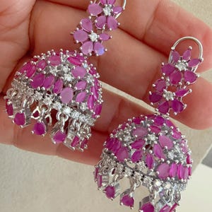 Peut inclure: Une paire de boucles d'oreilles en argent avec des pierres roses et blanches. Les boucles d'oreilles ont la forme d'une fleur avec un charme en forme de cloche.