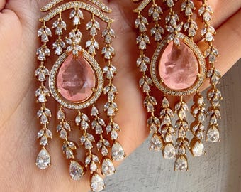 May include: Gold-toned chandelier earrings with a square top, intricate floral design, and a teardrop-shaped pink gemstone. The earrings feature dangling white gemstones and are held up by a gold hook.
