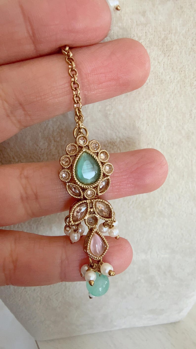 Pode incluir: Corrente de ouro com uma pedra verde em forma de l&aacute;grima cercada por strass transparentes, uma pedra rosa e p&eacute;rolas brancas.