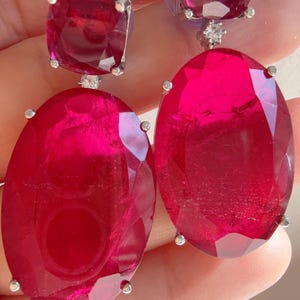 May include: A pair of ruby earrings. Each earring features a square-cut ruby at the top and a large oval-cut ruby below. The rubies are a vibrant red color and are set in a silver metal setting. Small diamonds are set between the ruby stones.