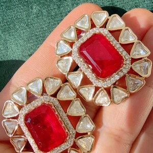 May include: A pair of gold-toned stud earrings. Each earring features a large, rectangular, red gemstone surrounded by a halo of small, clear, triangular stones. The earrings are set against a green background.