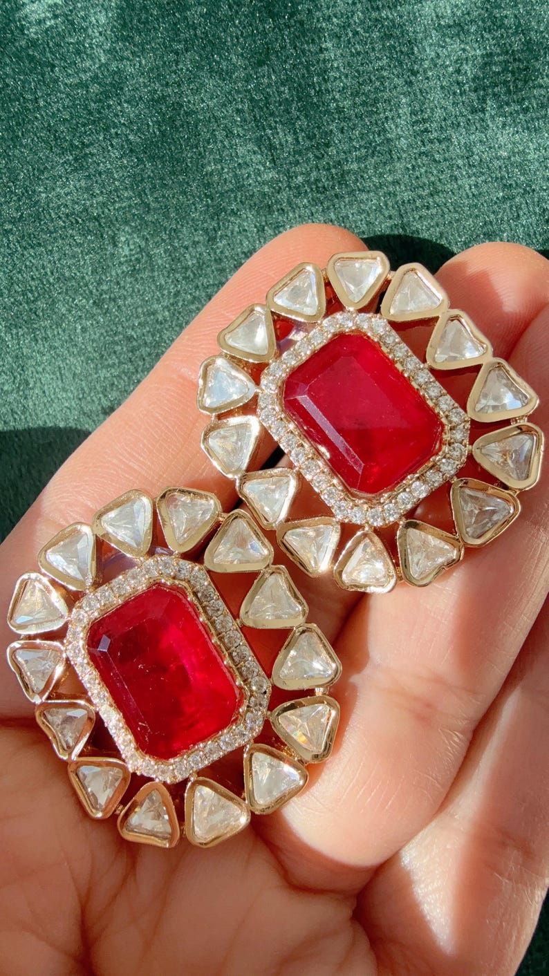 May include: A pair of gold-toned stud earrings. Each earring features a large, rectangular, red gemstone surrounded by a halo of small, clear, triangular stones. The earrings are set against a green background.