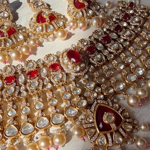 Puede incluir: Un conjunto de joyas de tono dorado con un collar y pendientes a juego. El collar y los pendientes est&aacute;n adornados con piedras preciosas rojas, cristales transparentes y perlas de imitaci&oacute;n. La joyer&iacute;a tiene un dise&ntilde;o vintage ornamentado.