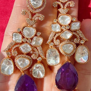 Puede incluir: Un par de pendientes dorados ornamentados con piedras preciosas claras y moradas. Los pendientes presentan un diseño complejo con múltiples piedras claras de forma irregular y una gran piedra morada en forma de lágrima en la parte inferior.
