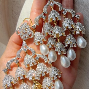 May include: Elegant chandelier earrings with a gold-tone setting, adorned with sparkling clear crystals and teardrop-shaped white pearls. The earrings have a cascading design, perfect for special occasions.