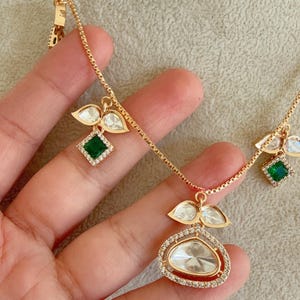 May include: A gold chain necklace with multiple pendants featuring clear and green gemstones. The pendants are shaped like leaves and squares.