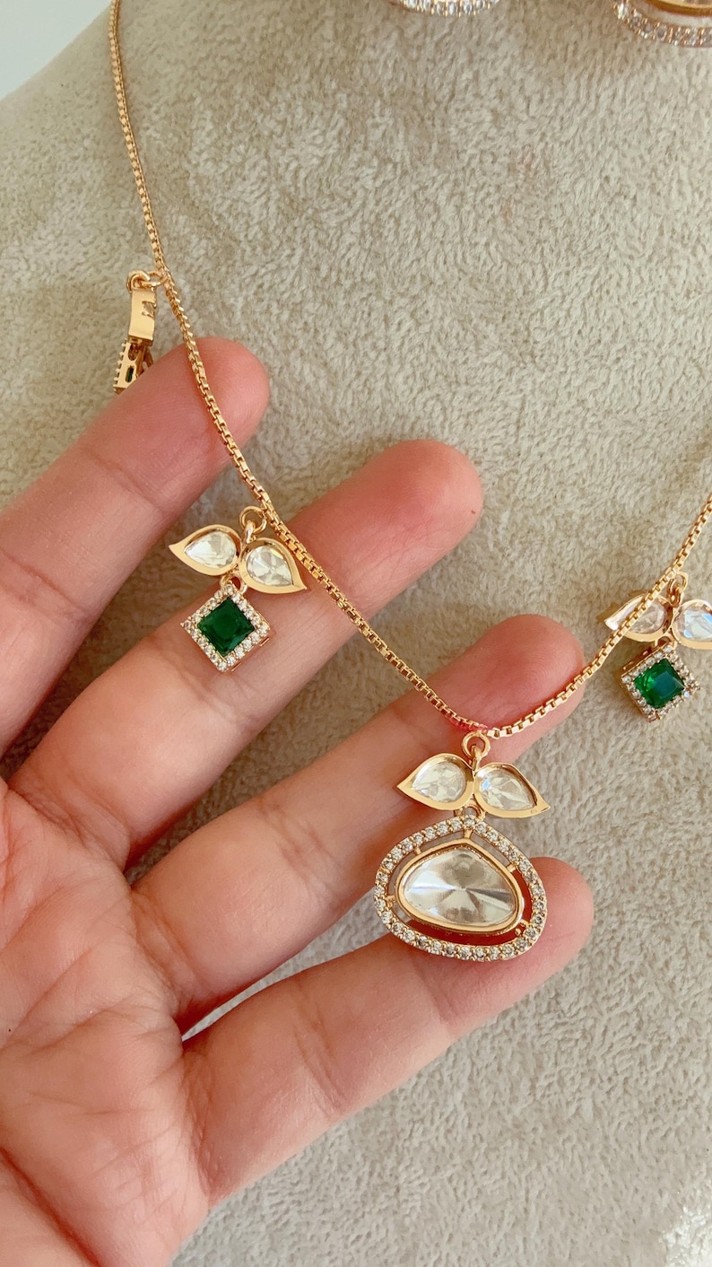 May include: A gold chain necklace with multiple pendants featuring clear and green gemstones. The pendants are shaped like leaves and squares.