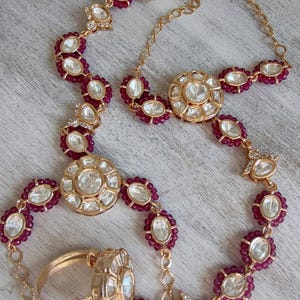 May include: A gold-toned jewelry set consisting of a necklace and two rings, featuring clear, flat-cut stones surrounded by small, deep pink beaded accents. The necklace includes a delicate chain with a lobster clasp and multiple circular stone settings.
