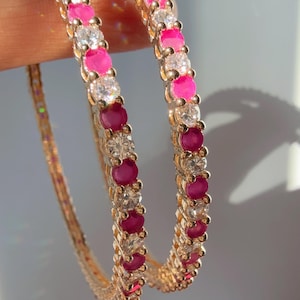 May include: Two gold-colored bangles with alternating pink and clear gemstones. The gemstones are round and set closely together, creating a continuous pattern around the bracelets. The background is a soft, neutral color.