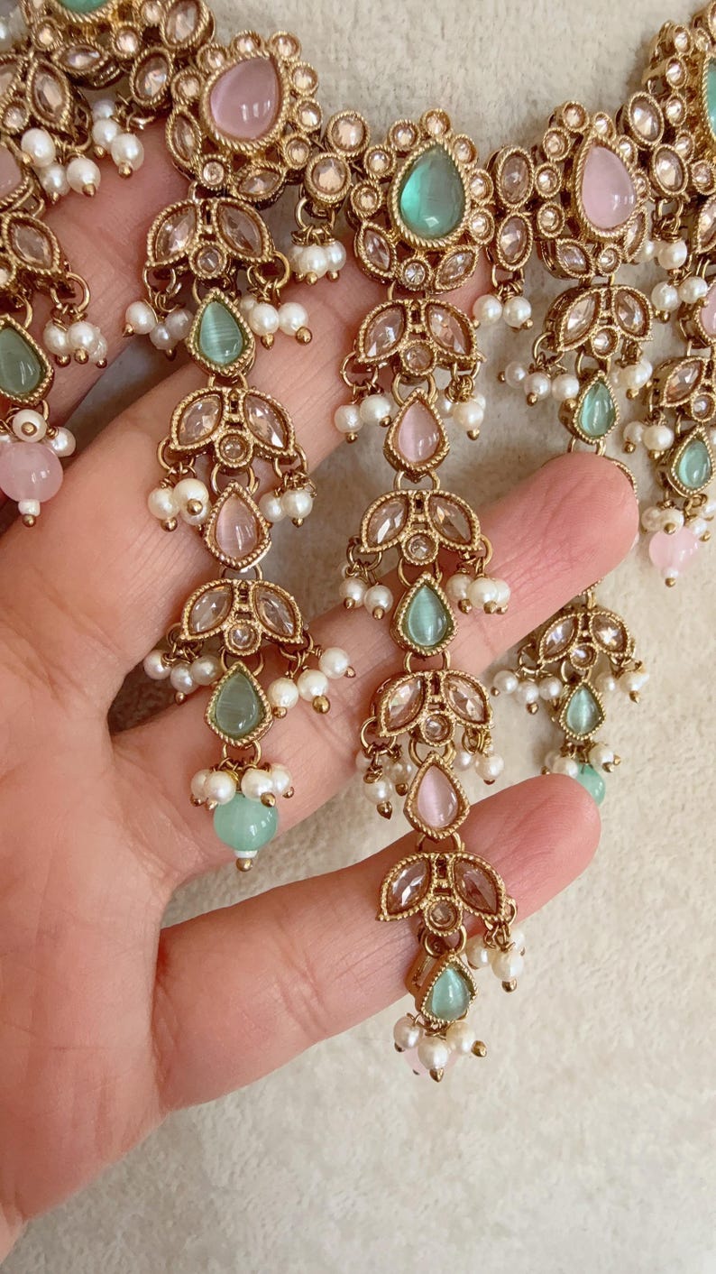 Pode incluir: Um colar de ouro com uma corrente delicada e v&aacute;rios pingentes. Os pingentes s&atilde;o adornados com pedras transparentes e de cor rosa e verde pastel. O colar &eacute; acentuado com pequenas p&eacute;rolas brancas.