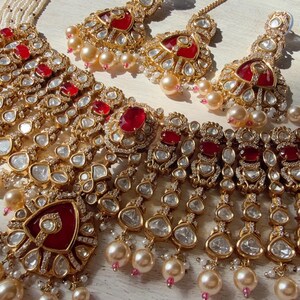 Puede incluir: Un conjunto de joyas dorado que incluye un collar y pendientes a juego. El collar y los pendientes est&aacute;n adornados con piedras preciosas rojas, cristales transparentes y perlas de imitaci&oacute;n. El conjunto se exhibe sobre una superficie clara, mostrando los intrincados detalles.