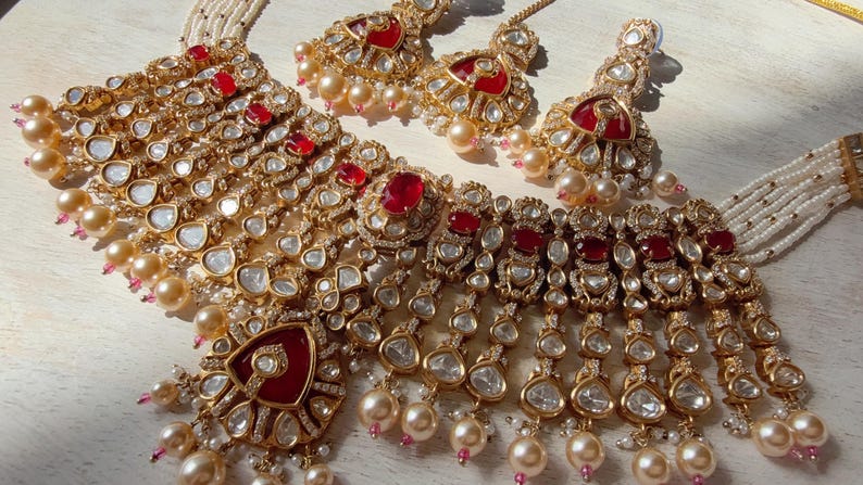 Puede incluir: Un conjunto de joyas dorado que incluye un collar y pendientes a juego. El collar y los pendientes est&aacute;n adornados con piedras preciosas rojas, cristales transparentes y perlas de imitaci&oacute;n. El conjunto se exhibe sobre una superficie clara, mostrando los intrincados detalles.