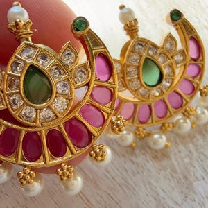 May include: Gold-toned earrings with a crescent shape, featuring a central green gemstone, surrounded by clear stones. The earrings have pink and red accents, with a pearl drop. A small green stone is at the top of the crescent.