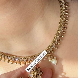 May include: A gold necklace with a teardrop-shaped design and white pearl accents. The necklace is paired with matching earrings featuring a flower design and white pearl accents. The text "Kundan Creations" is visible on the image.