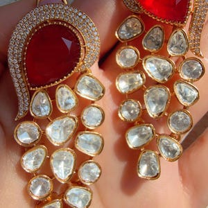 Peut inclure: Boucles d'oreilles dorées avec une grosse pierre rouge facettée en haut, entourée de petites pierres transparentes. En dessous, une cascade de pierres précieuses claires de forme irrégulière pend de la pierre du haut. Design élégant.