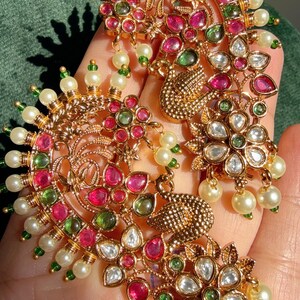 May include: Ornate gold-toned earrings with intricate floral and peacock designs. The earrings are adorned with red, green, and clear gemstones, along with small white pearls. The jewelry is set against a green background.