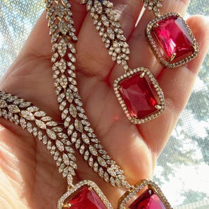 May include: Close-up of a gold-toned necklace and earrings set. The necklace and earrings feature a leaf-like design with small, clear gemstones. Each earring has a large, rectangular, red gemstone. The jewelry is displayed against a soft background.
