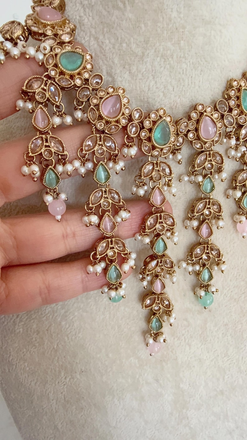 Pode incluir: Um colar de ouro com pedras rosa e verdes e detalhes de p&eacute;rolas. O colar tem um design em camadas com v&aacute;rias fileiras de p&eacute;rolas e pedras.