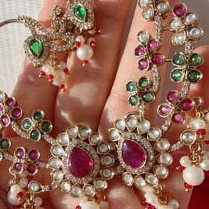 May include: Gold-toned jewelry set featuring floral and teardrop designs. The set includes earrings and a necklace, adorned with red, green, and white gemstones, and small white pearls. The intricate design is set against a light background.