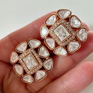 May include: A pair of gold-tone earrings with a square-shaped center stone surrounded by smaller stones. The earrings are set in a floral design.