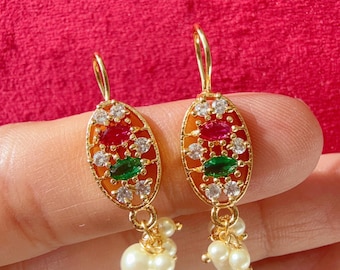 Pode incluir: Brincos dourados com pingentes ovais. Cada pingente apresenta pedras preciosas vermelhas, verdes e transparentes, com um aglomerado de pequenas pérolas penduradas abaixo. Os brincos estão em um fundo vermelho.