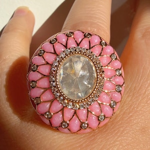 May include: A large, pink statement ring with a central, clear gemstone. The ring features a floral design with small, sparkling accents and a gold-coloured band. The ring is a bold piece of jewellery.