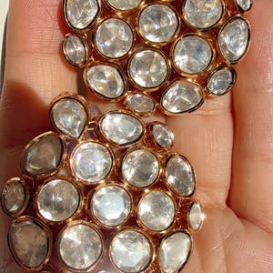 May include: Two gold-toned, cluster earrings, each featuring numerous clear, faceted gemstones. The gemstones are oval and round, set closely together. The earrings are displayed against a light background.