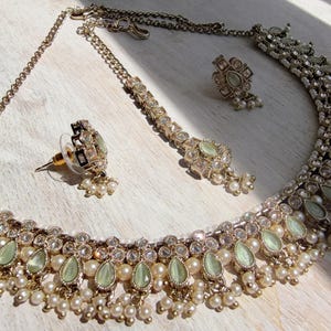 May include: A jewelry set consisting of a necklace, two earrings, and a maang tikka. The pieces feature light green teardrop-shaped stones, clear crystals, and small white pearls set in a metallic frame with a delicate chain and post backings.