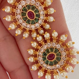May include: A pair of gold earrings with a circular design. The earrings are adorned with white pearls, clear rhinestones, and green and red gemstones.