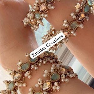 Può includere: Primo piano di braccialetti dorati adornati con perle e pietre preziose. I braccialetti presentano un design con pietre verdi e color pesca a forma di goccia, circondate da piccole perle bianche. Il testo "Kundan Creations" è visibile.