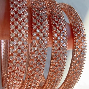 Op de afbeelding: Roségouden armbanden bezet met sprankelende, heldere edelstenen. De armbanden zijn op elkaar gestapeld, wat hun ingewikkelde ontwerp en reflecterende kwaliteiten benadrukt. Perfect om een vleugje elegantie toe te voegen.