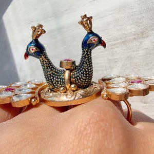 Peut inclure: Bague dorée ornée de deux paons aux corps bleus et noirs, aux becs rouges et aux couronnes dorées. La bague est ornée de pierres précieuses blanches et rouges, créant un bijou unique et orné.