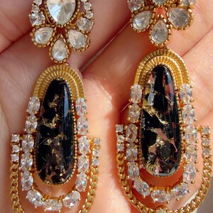 May include: A pair of gold-toned drop earrings. Each earring features a large, oval black stone with gold flecks, surrounded by clear, sparkling crystals. The earrings have a chain-like design and are adorned with additional crystals.