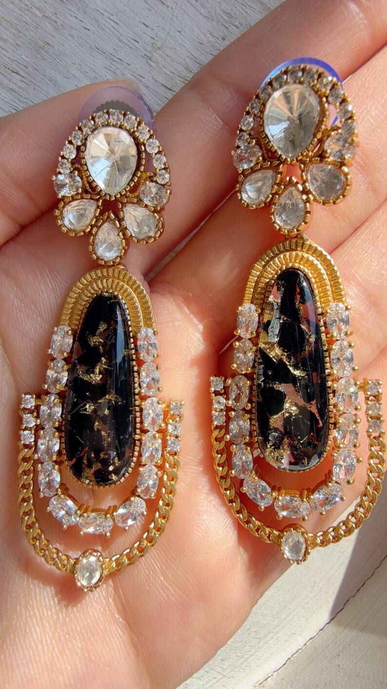 May include: A pair of gold-toned drop earrings. Each earring features a large, oval black stone with gold flecks, surrounded by clear, sparkling crystals. The earrings have a chain-like design and are adorned with additional crystals.