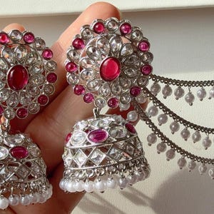 May include: Silver-toned earrings with a floral design, featuring a large, round, red gemstone in the center, surrounded by smaller clear stones. The earrings have a bell-shaped bottom with more red gemstones and white pearls.