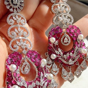 Peut inclure: Boucles d'oreilles argentées élégantes avec un motif floral. Elles présentent un ensemble de petites pierres précieuses roses vives et de cristaux clairs étincelants. De petites perles rondes sont intégrées, ajoutant une touche de sophistication.