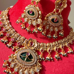 Könnte beinhalten: Ein goldfarbenes Schmuckset mit Halskette und passenden Ohrringen. Das Set umfasst eine Halskette mit einem großen Anhänger und Ohrringe, die alle mit roten und grünen Edelsteinen, weißen Perlen und aufwendigen Details verziert sind. Der Schmuck wird auf rotem Untergrund präsentiert.
