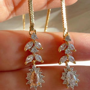 May include: Gold-toned drop earrings with a delicate design. Each earring features a chain, a cluster of clear, leaf-shaped stones, and a pear-shaped, peach-colored stone surrounded by smaller clear stones. The earrings are held up by a hand.