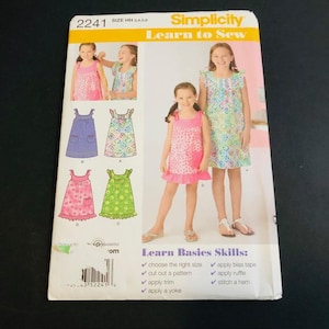 May include: Simplicity Learn to Sew pattern 2241 for girls' dresses, sizes HH 3, 4, 5, and 6. The pattern features illustrations of various dress designs, including a pink polka dot dress with a ruffle hem and a green floral dress. The package also lists basic sewing skills.