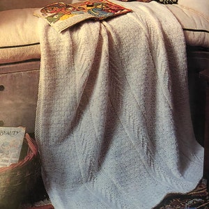 May include: A white knitted blanket with a subtle pattern, draped over a light brown couch with a book and a teddy bear on top. The blanket is folded at the bottom and the edges are visible.
