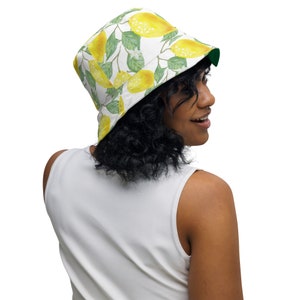 May include: Reversible bucket hat featuring a lemon pattern on a white background. The hat has a green underside. The lemons are yellow with green leaves and white flowers. The hat is worn by a person.