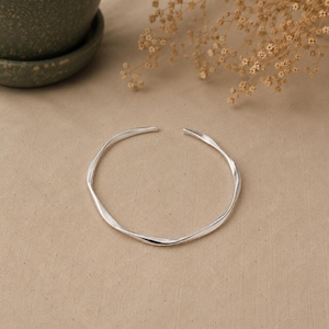 Peut inclure: Un bracelet ouvert de couleur argentée avec une surface lisse, ondulée et sinueuse. Le bracelet présente un design circulaire et flexible avec une petite ouverture sur le dessus, mettant en valeur une finition métallique polie et réfléchissante.