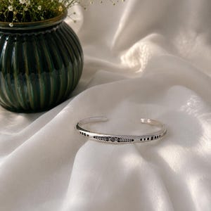 Peut inclure: Un bracelet manchette en argent orné d'un motif central de feuilles gravées, entouré de petits motifs circulaires. Le bracelet métallique présente une ouverture et une finition polie avec des détails oxydés sombres dans les gravures.