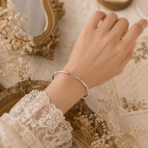 Peut inclure: Un bracelet ouvert de couleur argentée doté d'une finition lisse et polie avec de petites extrémités sphériques arrondies. Ce bracelet est conçu comme une bande métallique simple et épurée qui s'enroule partiellement autour du poignet.