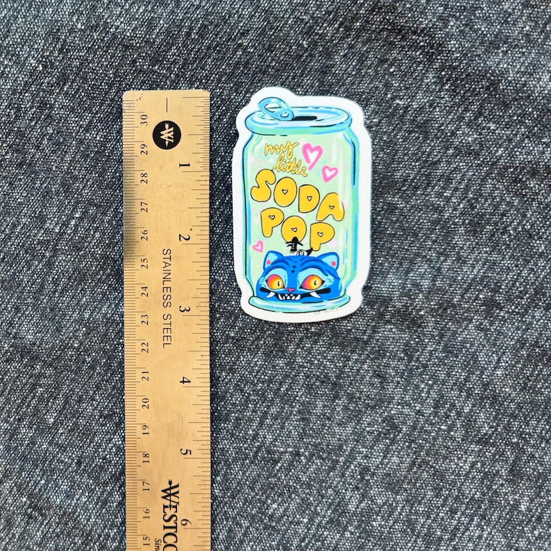 May include: A colorful sticker featuring a cartoon soda can design. The can is light blue with the words "my little SODA POP" in yellow. A blue cat with orange eyes is at the bottom. The sticker measures approximately 3 inches in height.