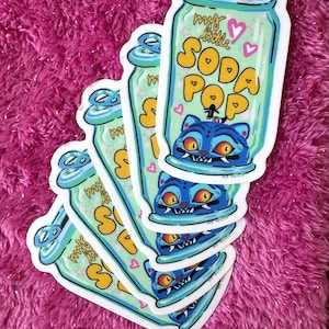 May include: A stack of stickers featuring a soda can design with the text "my little SODA POP". The can is light green with blue accents and a blue cat illustration. The background is a vibrant pink.