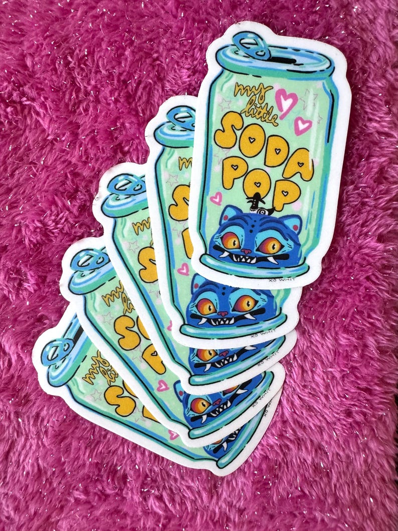 May include: A stack of stickers featuring a soda can design with the text "my little SODA POP". The can is light green with blue accents and a blue cat illustration. The background is a vibrant pink.